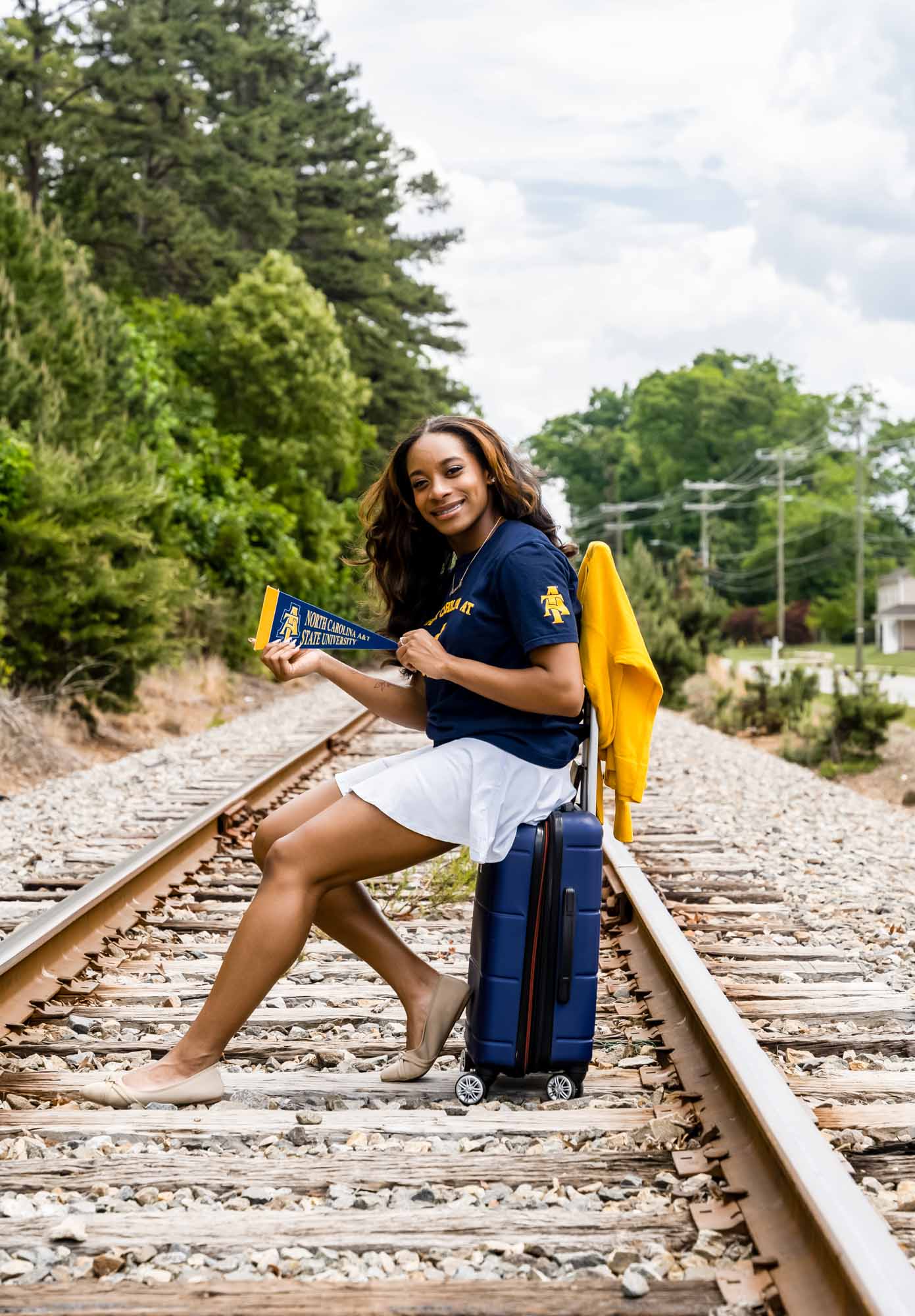 High school senior sitting on luggage on train tracks wearing future college attire