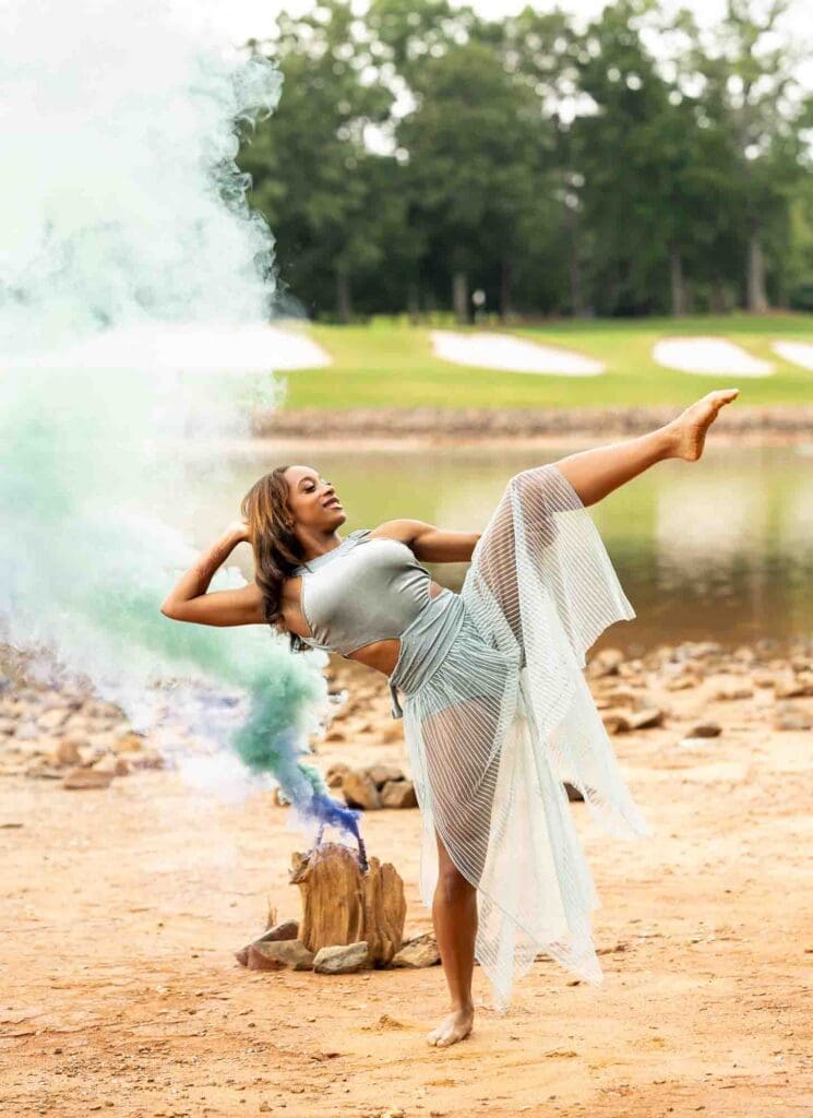 High school senior dancing on the waterfront with vibrant blue and purple smoke bombs during a Charlotte Senior Signature Experience at Jetton Park.